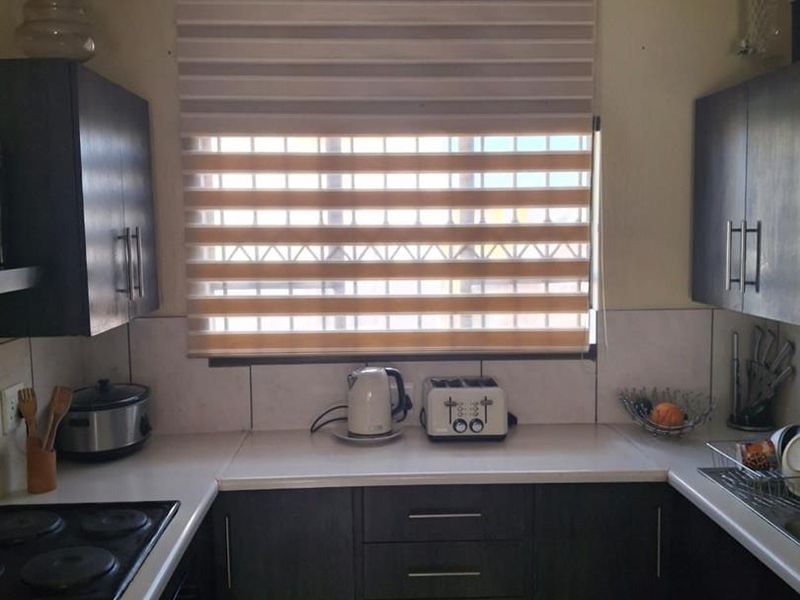 A compact kitchen featuring dark cabinetry, white countertops, an electric hob, and a tiled splashback, illuminated by a window with modern roller blinds.