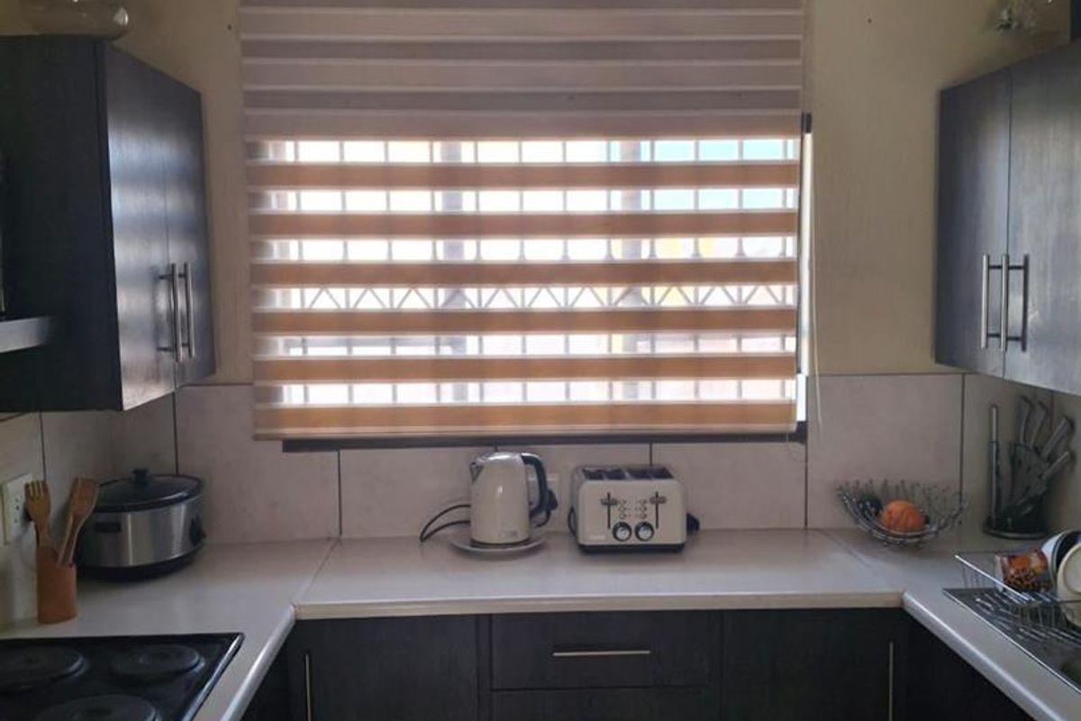 A compact kitchen featuring dark cabinetry, white countertops, an electric hob, and a tiled splashback, illuminated by a window with modern roller blinds.