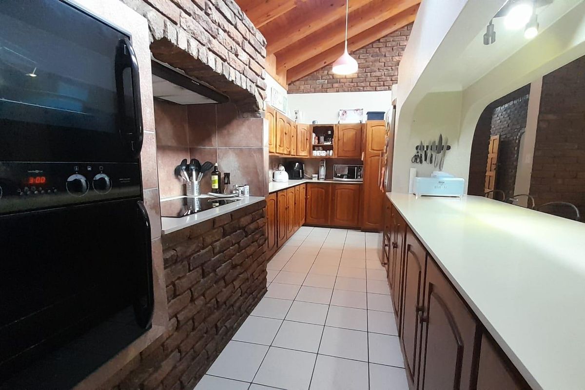 Spacious kitchen featuring rustic brick accents, wooden cabinetry, built-in oven, hob, and a long breakfast counter under a vaulted timber ceiling.