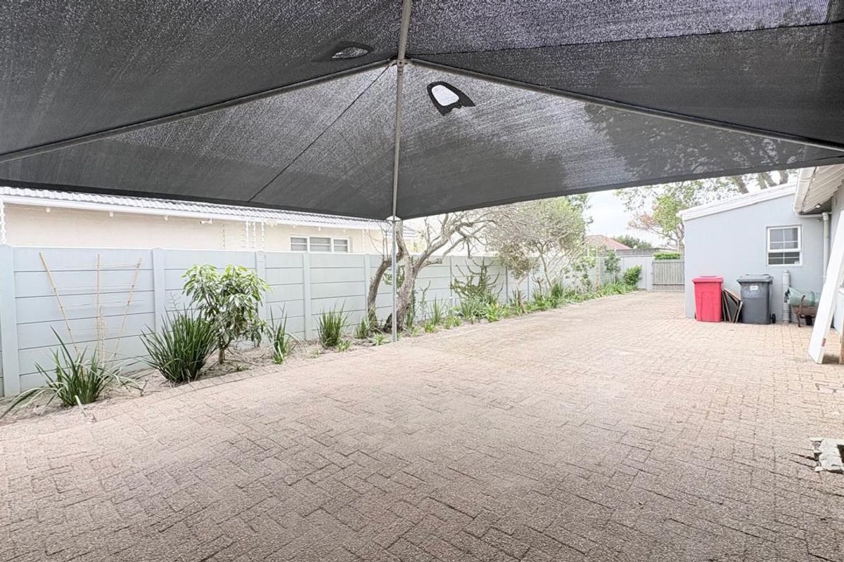 A paved rear outdoor area featuring a large shade sail, perimeter precast walling, and established garden beds.