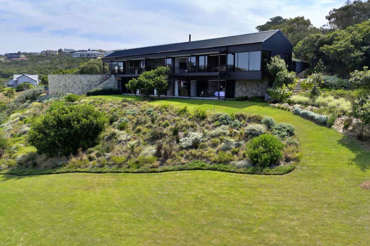A modern, architecturally designed two-story home featuring dark metal cladding, expansive glass facades, and stone retaining walls, set within a landscaped garden.