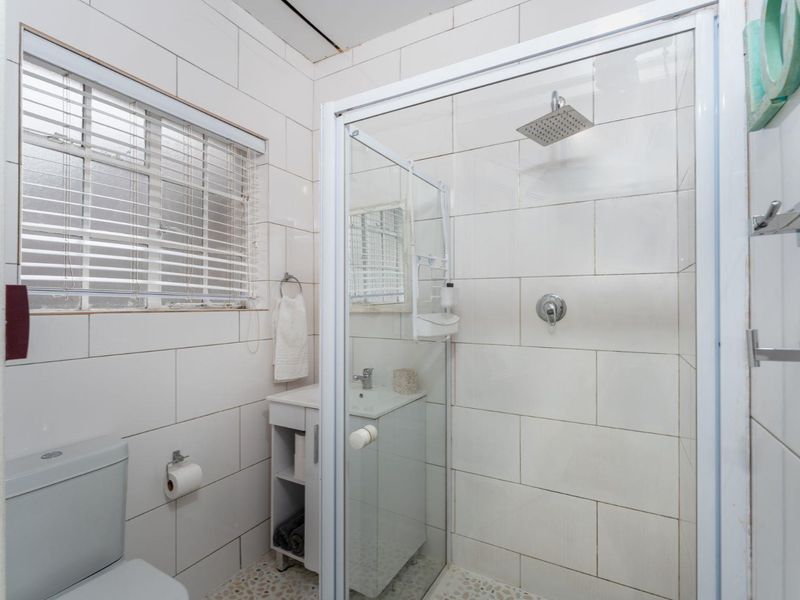 Compact bathroom featuring a glass-enclosed shower with a rain shower head, a vanity basin, and a toilet, finished with white wall tiles.