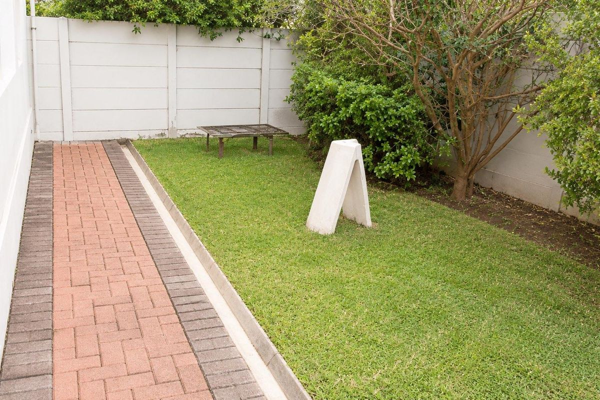 A private rear garden area featuring a paved walkway, a well-maintained lawn, and a secure precast concrete boundary wall.