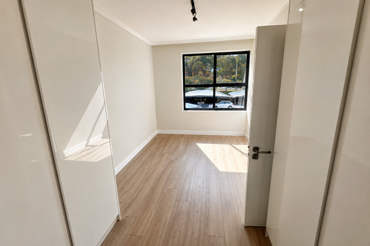 A bright, empty bedroom featuring modern laminate flooring, built-in cupboards with high-gloss finish, and a large window with black frames.