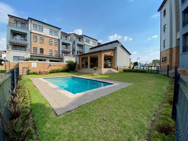 Communal outdoor area featuring a swimming pool, paved surround, landscaped lawn, and a covered patio area within an apartment complex.