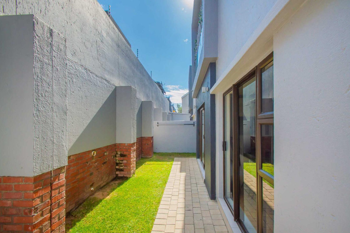 A side garden area featuring a paved walkway, a small lawn, and a boundary wall with integrated electric fencing, adjacent to the main building structure.