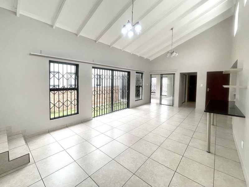Spacious living area featuring vaulted ceilings with exposed beams, neutral tiled flooring, and large windows with security gates, offering a bright and open layout.
