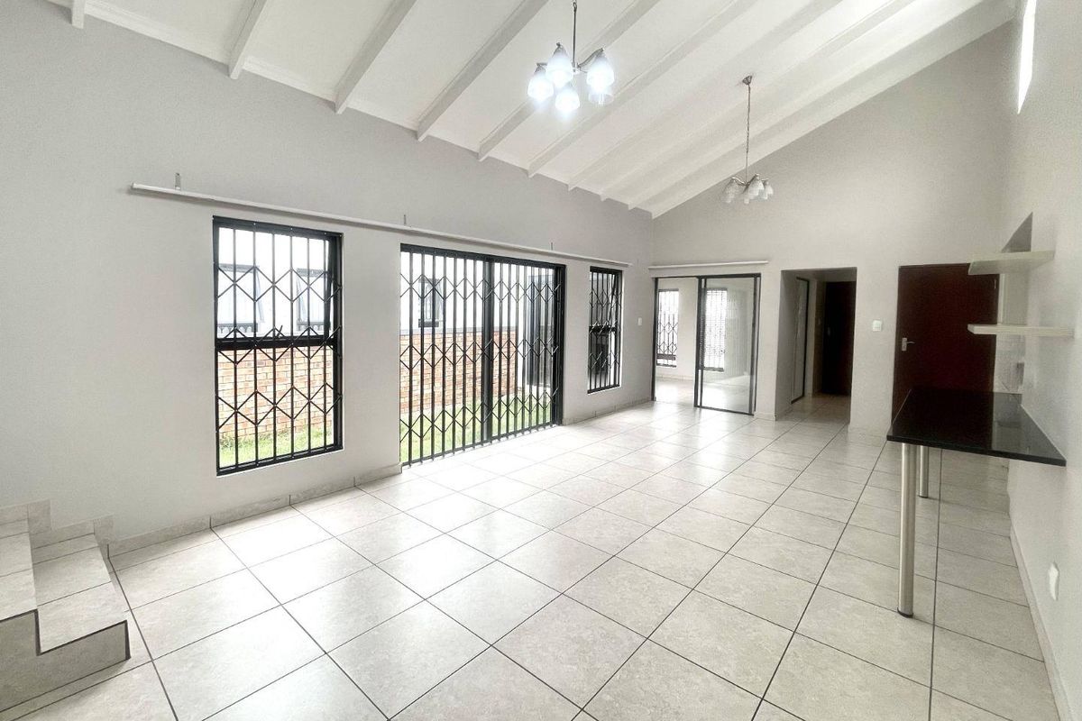 Spacious living area featuring vaulted ceilings with exposed beams, neutral tiled flooring, and large windows with security gates, offering a bright and open layout.