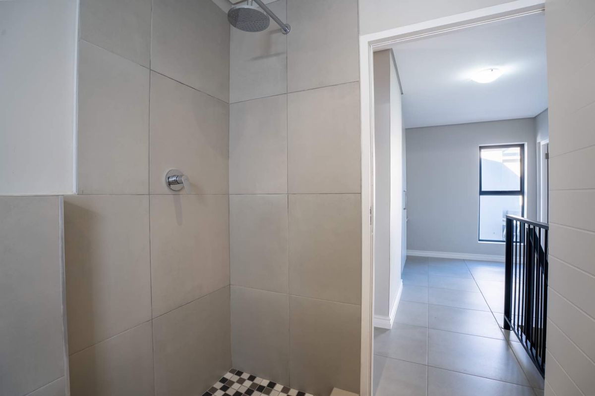 A modern bathroom shower area featuring neutral large-format wall tiles and a contrasting mosaic floor, adjacent to a bright hallway with staircase access.