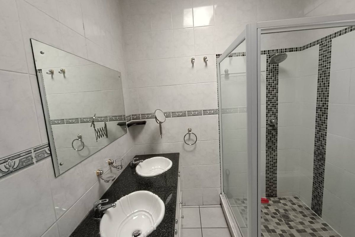 A functional bathroom featuring a double vanity with granite countertop, wall-mounted mirror, and a glass-enclosed shower with decorative mosaic tile accents.