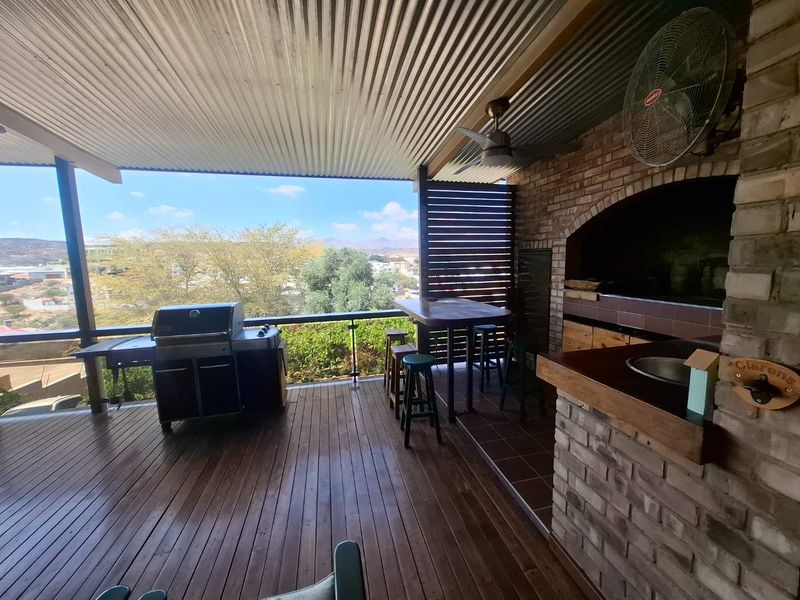Covered outdoor patio featuring timber decking, a built-in brick braai, prep area with sink, and elevated views of the surrounding landscape.