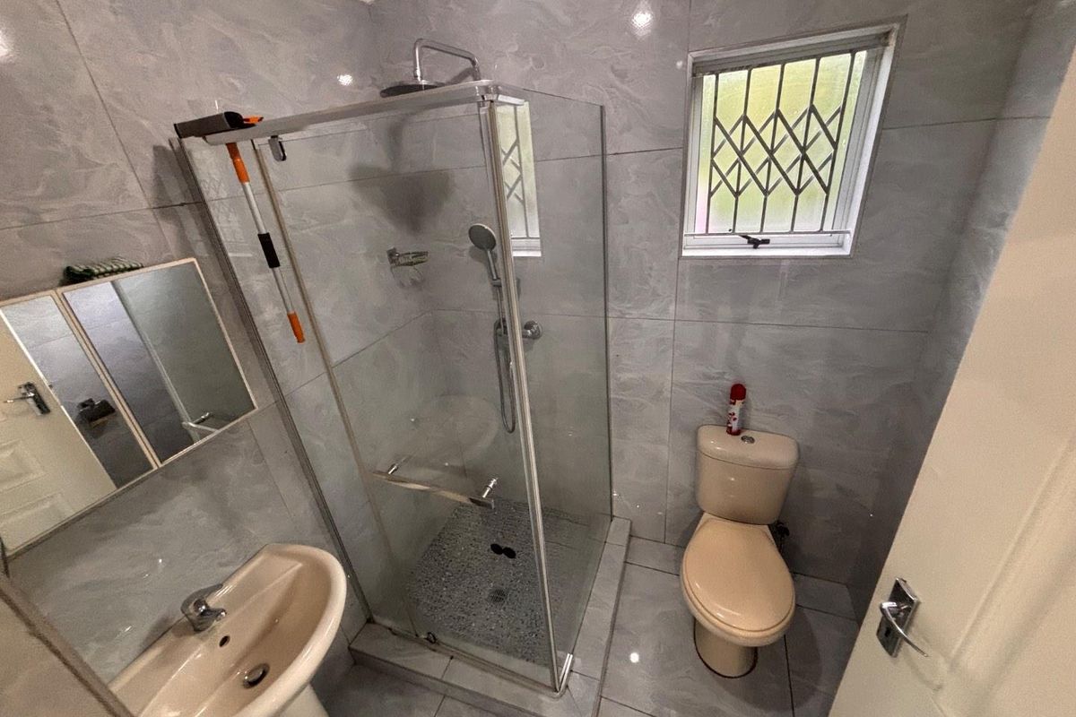 Compact bathroom featuring a glass-enclosed shower, pedestal basin, and toilet with full-height wall tiling and a security-grilled window.