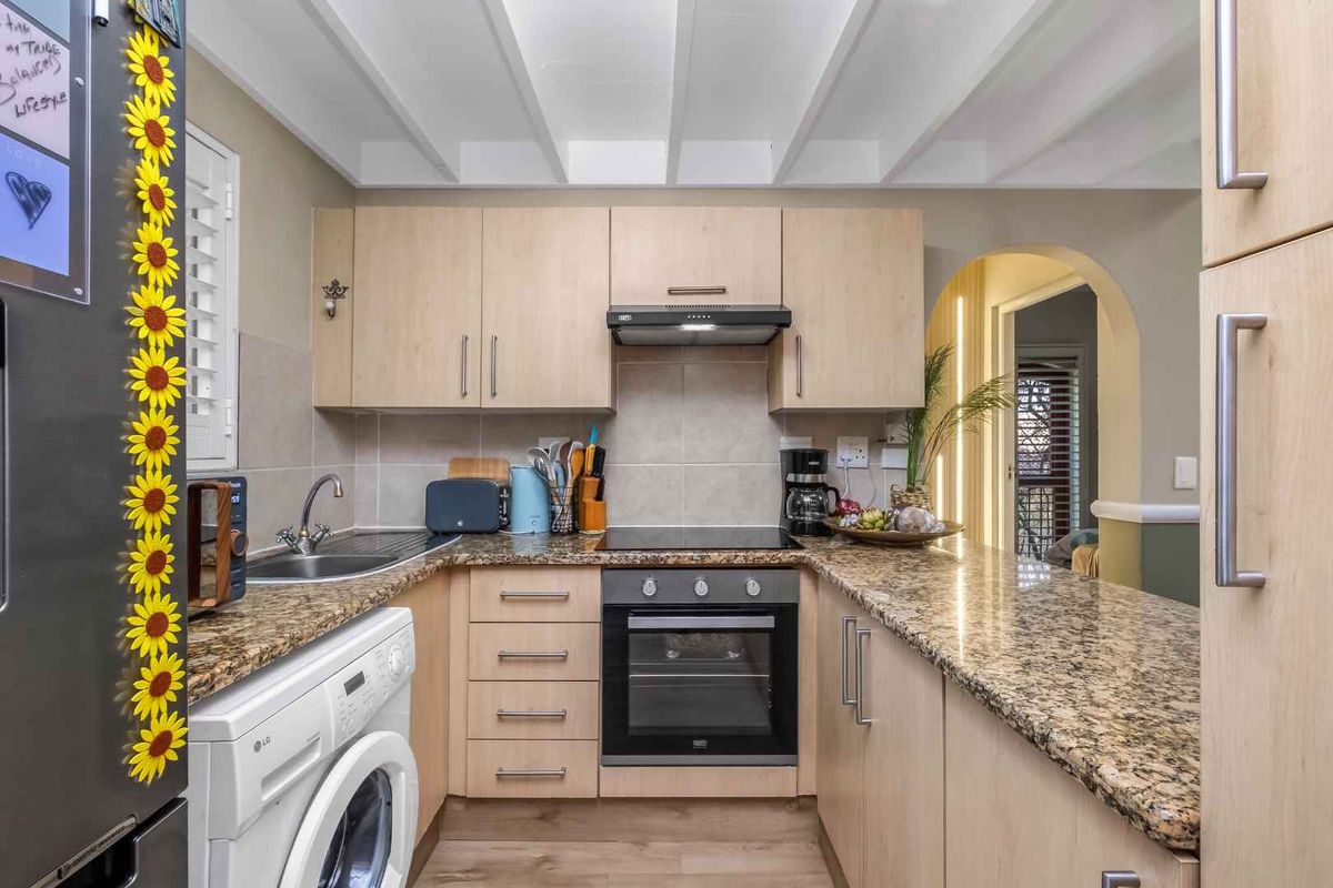 Compact kitchen featuring light wood cabinetry, granite countertops, built-in oven, electric hob, and an integrated laundry space, finished with an exposed beam ceiling.