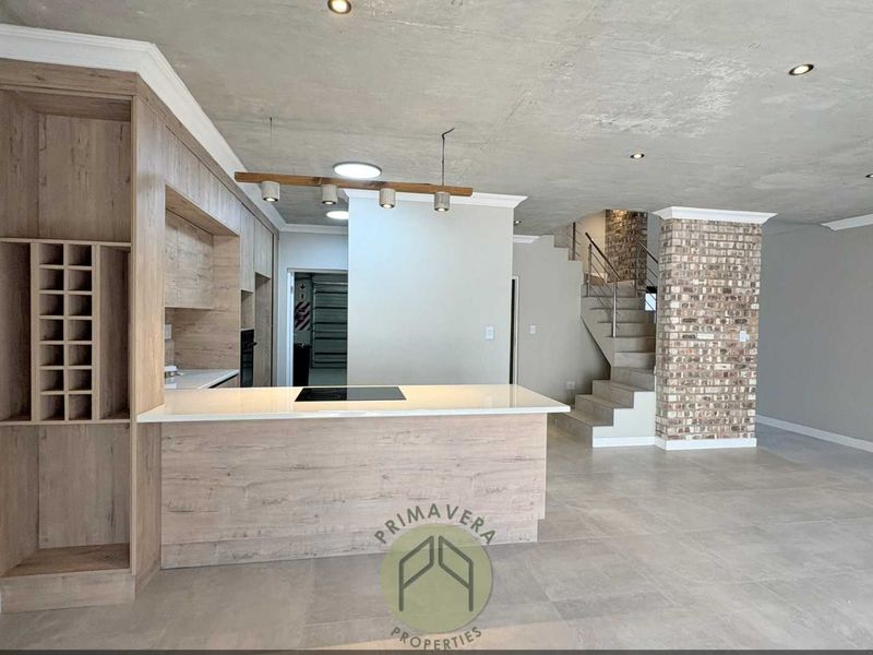 Modern open-plan living area featuring a sleek kitchen with stone countertops, induction hob, exposed concrete ceilings, and a striking brick feature pillar with staircase.