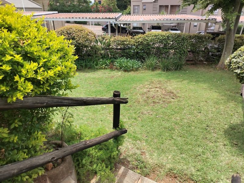 A private garden area featuring a lawn, established shrubbery, and a timber fence, with a communal carport structure visible in the background.