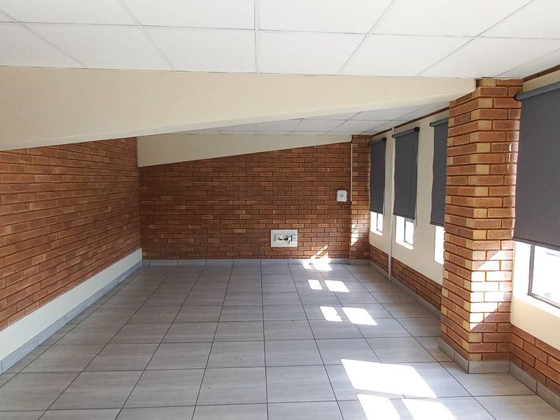 An open-plan industrial office space featuring exposed brick walls, tiled flooring, suspended ceiling tiles, and fitted roller blinds on windows.