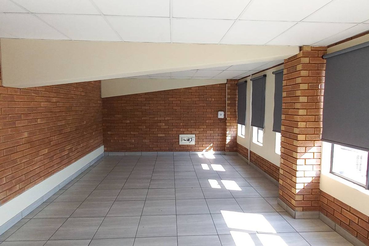 An open-plan industrial office space featuring exposed brick walls, tiled flooring, suspended ceiling tiles, and fitted roller blinds on windows.
