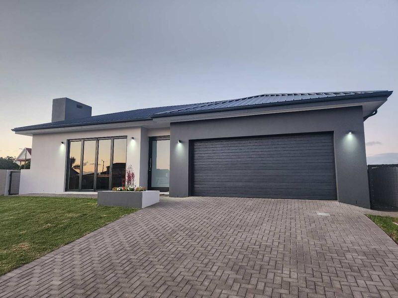 Modern single-story townhouse exterior featuring a paved driveway, integrated garage, contemporary metal roof, and large glass sliding doors with exterior lighting.