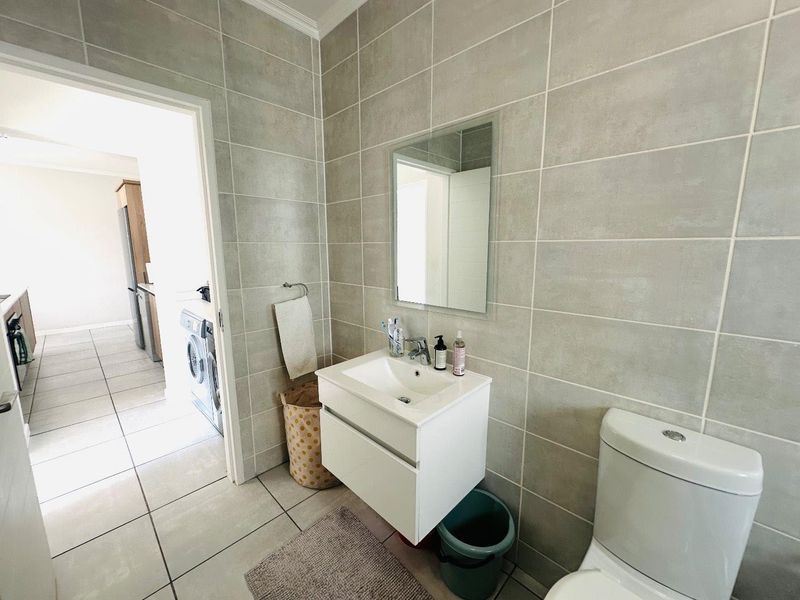 A modern bathroom featuring a wall-mounted vanity, ceramic basin, and neutral wall-to-floor tiling, with an adjacent view into the laundry area.