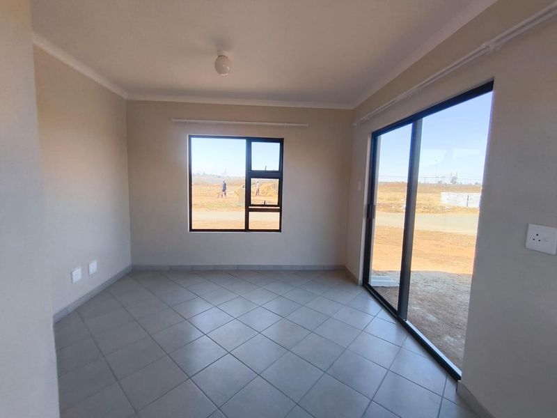 A bright, empty living area featuring neutral tiled flooring, large aluminium-framed windows, and a sliding glass door providing access to the exterior.