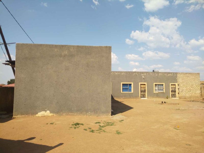 Rear view of a single-story house featuring a cement plaster finish, security bars on windows, and an expansive, unlandscaped dirt yard.