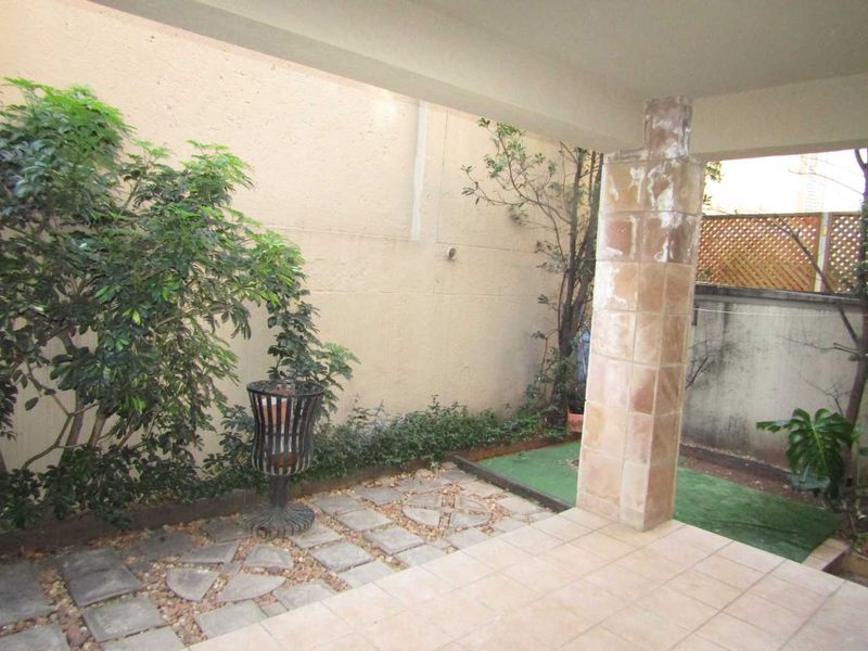 A covered patio area featuring tiled flooring and a tiled support column, leading to a small paved and landscaped garden space.
