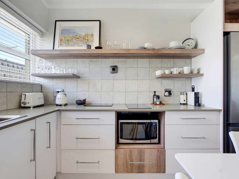 Modern kitchen featuring clean white cabinetry, stone countertops, a built-in microwave, and stylish floating timber shelving with a tiled backsplash.