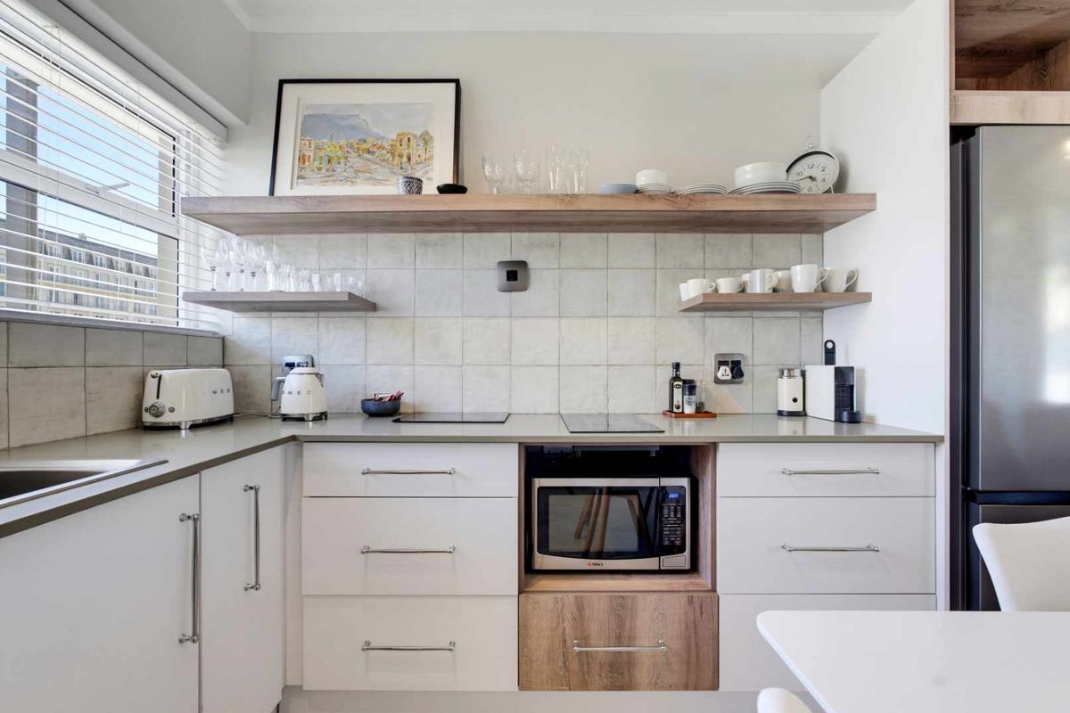 Modern kitchen featuring clean white cabinetry, stone countertops, a built-in microwave, and stylish floating timber shelving with a tiled backsplash.