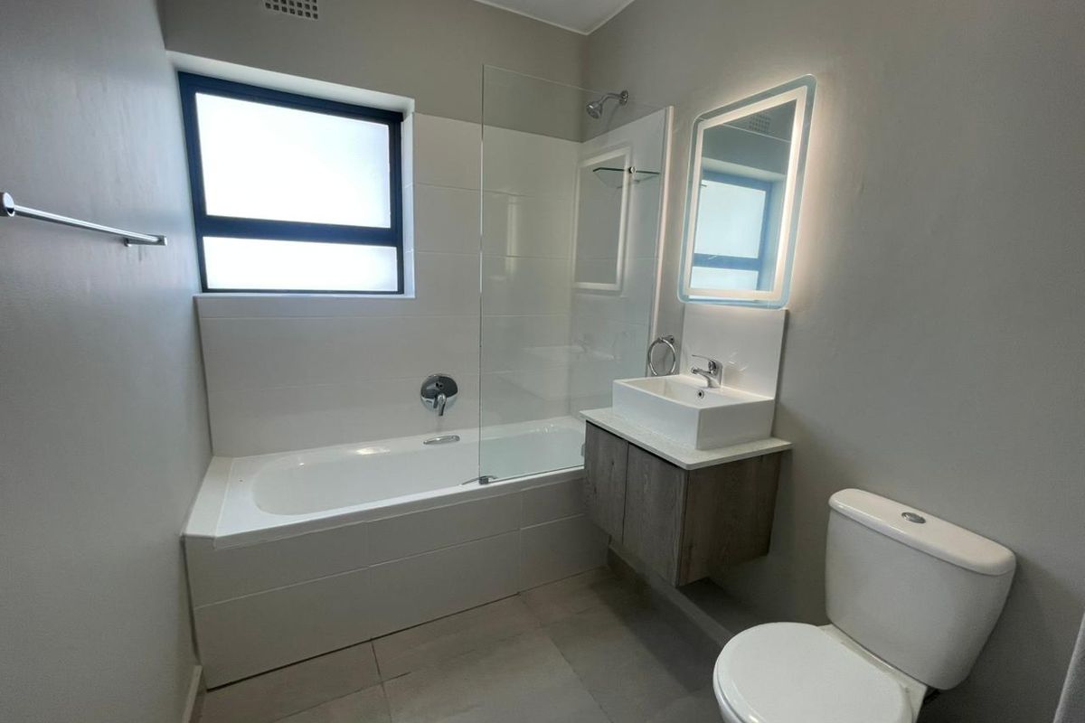 Modern bathroom featuring a built-in bathtub with glass shower screen, wall-mounted vanity with basin, illuminated mirror, and a standard toilet.