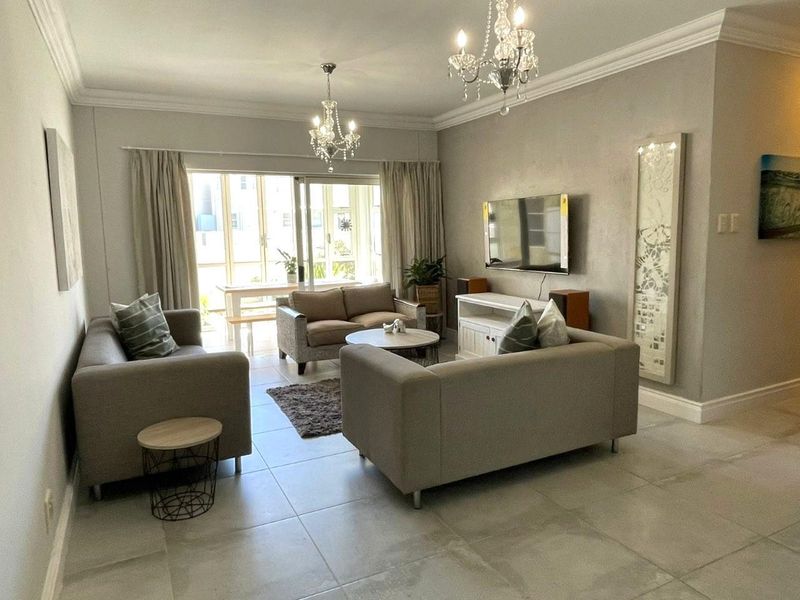 A well-maintained living room featuring large format floor tiles, elegant crown molding, dual crystal chandeliers, and a sliding glass door leading to an outdoor area.