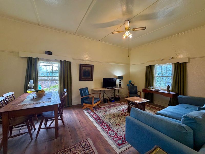 Open plan living and dining area featuring original timber flooring, ceiling fan, and security bars on windows, showing signs of wear and wall cracking.