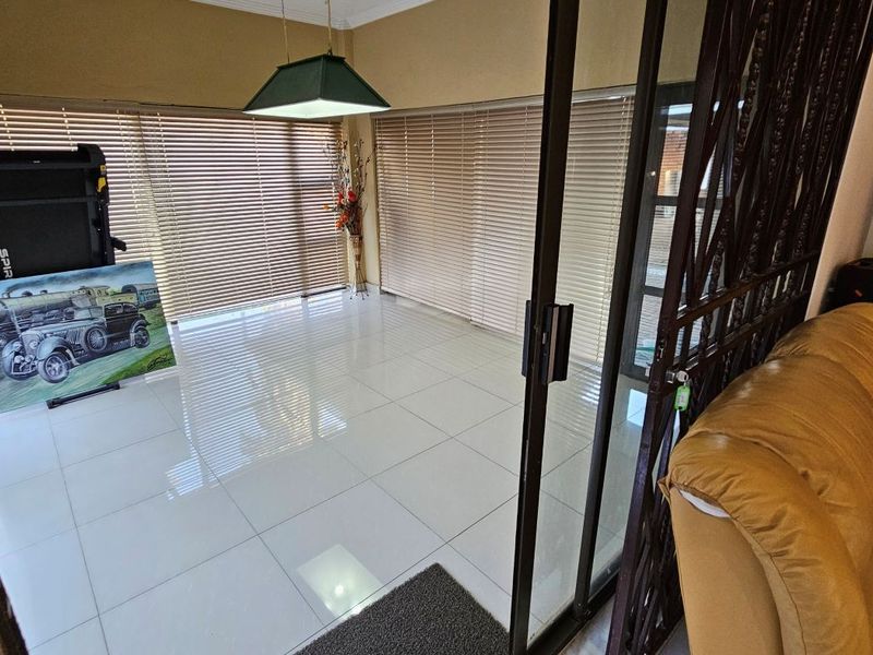 A bright living area featuring large windows with venetian blinds, polished tiled flooring, and sliding glass doors with a security gate.