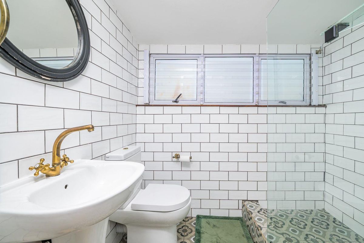 A bright bathroom featuring white subway wall tiles, patterned floor tiles, a pedestal basin with brass tapware, and a glass-enclosed shower.