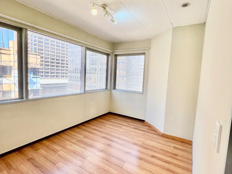 A bright, empty bedroom featuring laminate flooring, large windows with city views, and a suspended ceiling with integrated lighting and air conditioning.