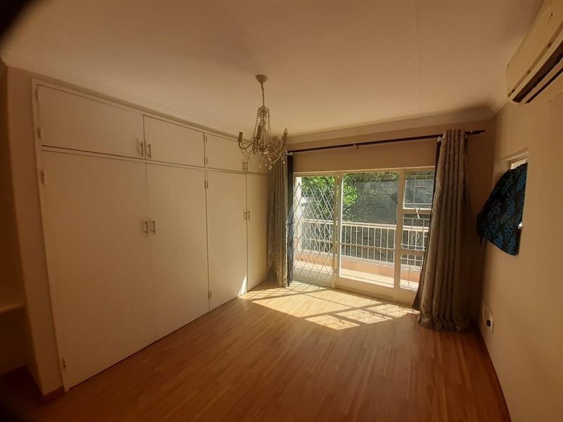 A bright bedroom featuring extensive built-in cupboards, laminate flooring, an air conditioning unit, and direct access to a private balcony through a sliding door.