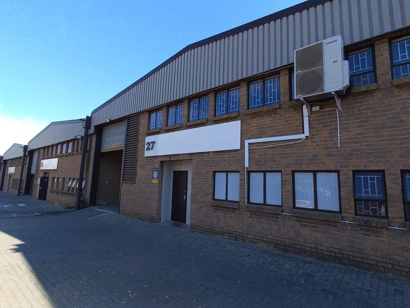 Exterior view of an industrial warehouse unit featuring brick facade, roller shutter door, and mounted air conditioning unit.