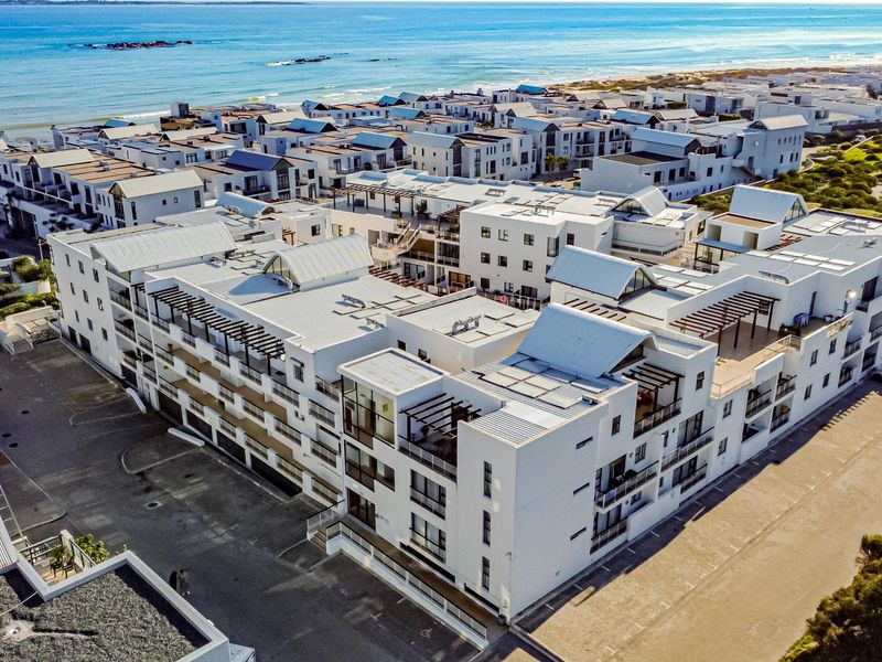 Aerial view of a modern coastal apartment complex featuring white facades, metal roofing, and balconies, situated near the ocean.