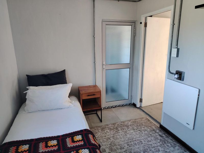 A compact bedroom featuring tiled flooring, exposed electrical conduit, and a wall-mounted panel heater, with direct access to an adjacent space.