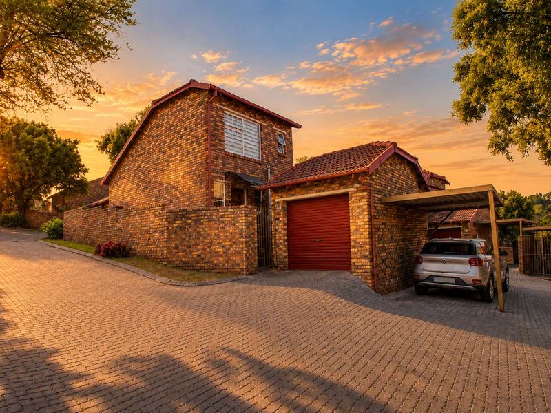 A well-maintained face brick townhouse featuring a single garage, attached carport, and paved driveway, set within a neat residential complex at sunset.