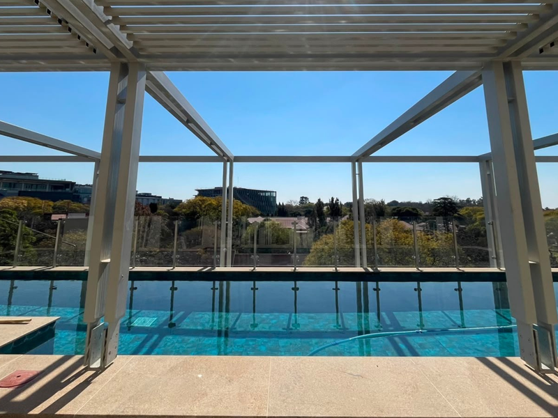 A modern rooftop swimming pool area featuring a sleek metal pergola, glass balustrades, and expansive views of the surrounding neighborhood.