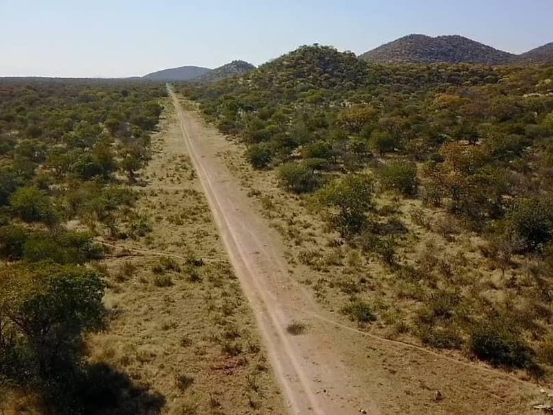 An aerial view of a long dirt road stretching through a vast, natural farm landscape with rolling hills and indigenous vegetation.