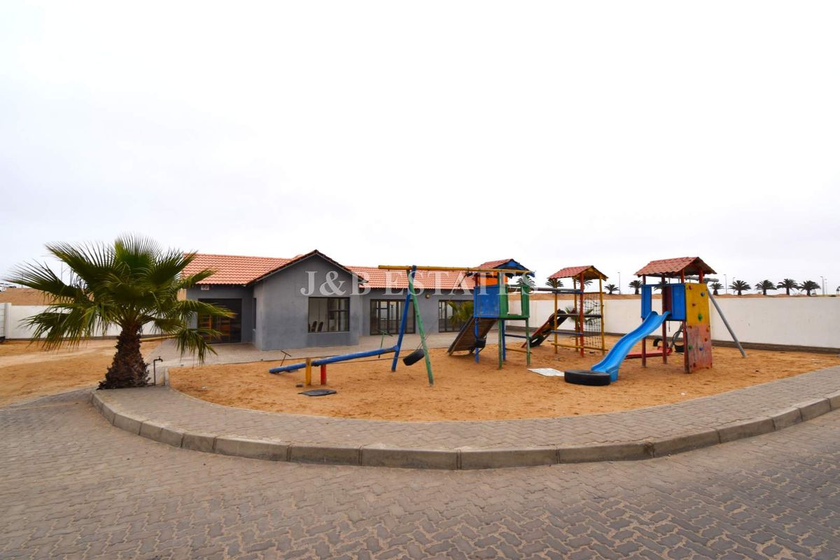 Exterior view of a residential property featuring a communal playground area with sand surface and a single-story building with a tiled roof.