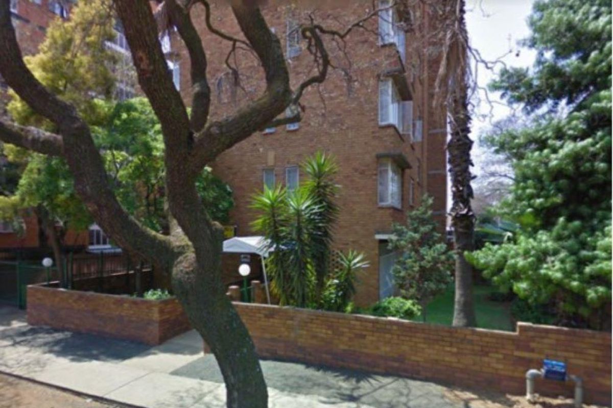 Exterior view of a brick apartment building facade featuring a low brick boundary wall, mature trees, and landscaped garden area.