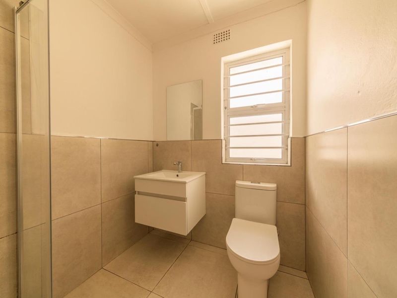 A clean, modern bathroom featuring a wall-mounted vanity, a toilet, and a glass-enclosed shower with neutral floor-to-ceiling tiling.