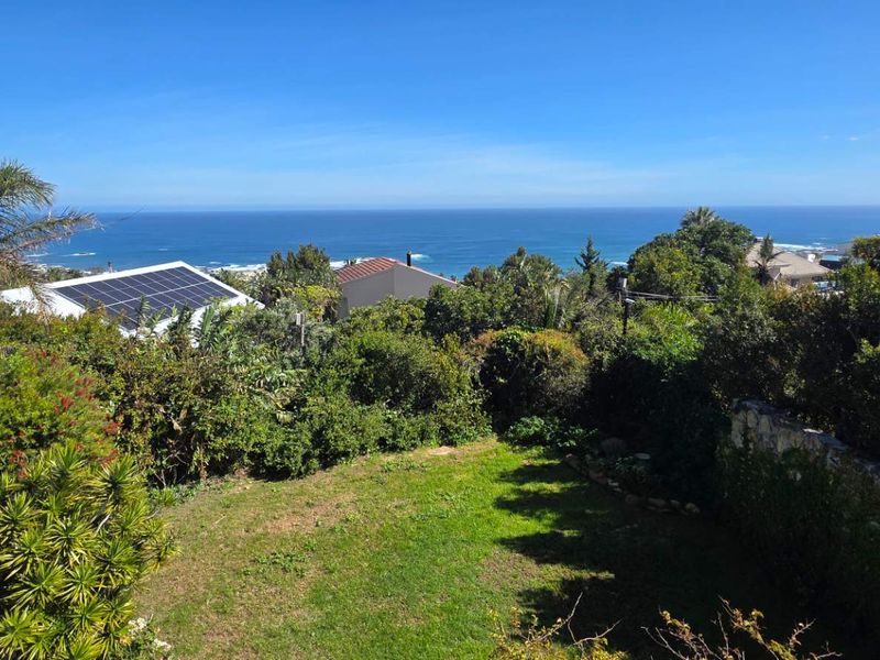 Elevated view overlooking a lush garden with a stone retaining wall, featuring a clear ocean horizon and neighbouring properties with solar installations.