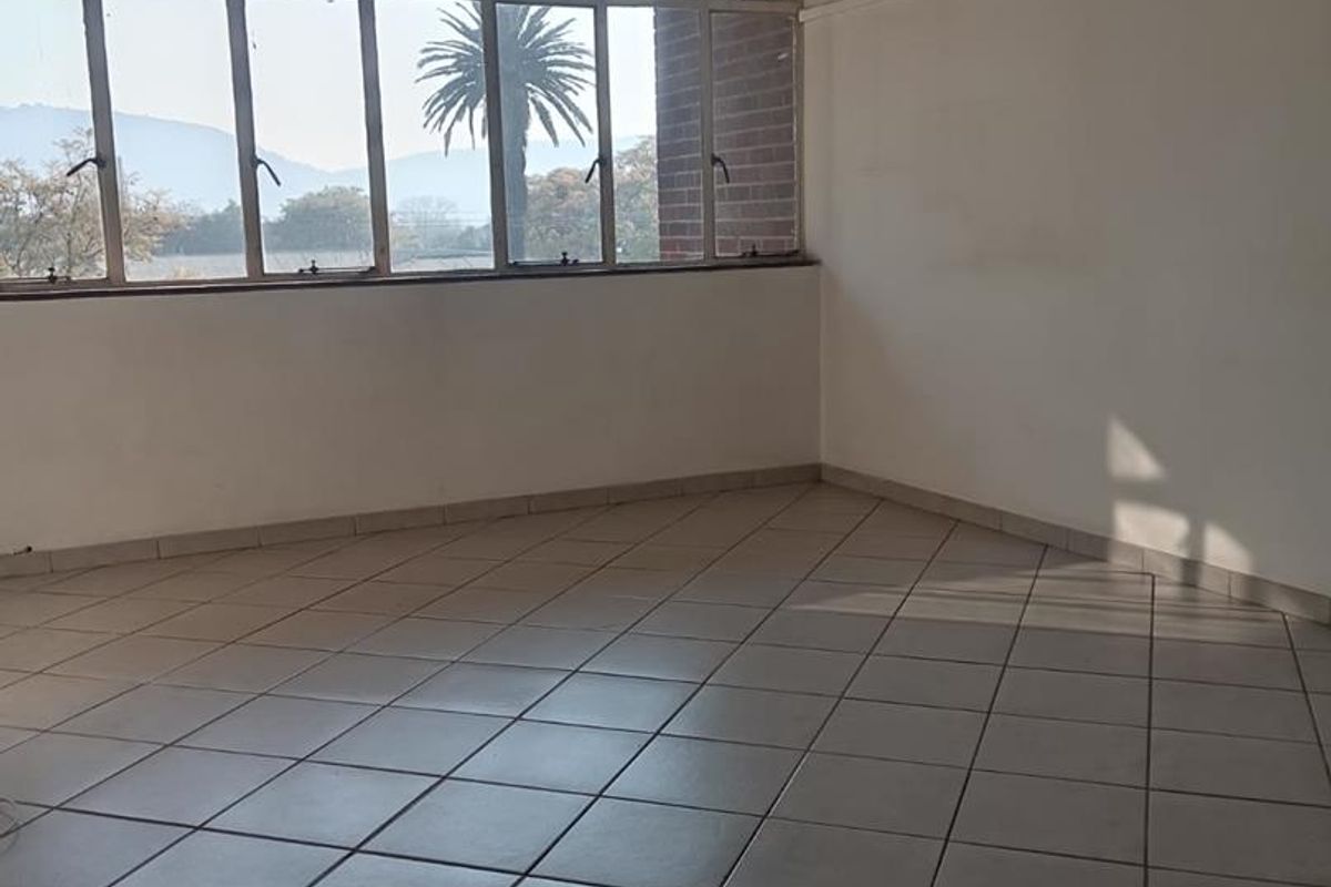 An empty, sunlit living area featuring large multi-pane windows, ceramic tiled flooring, and a feature brick wall section.