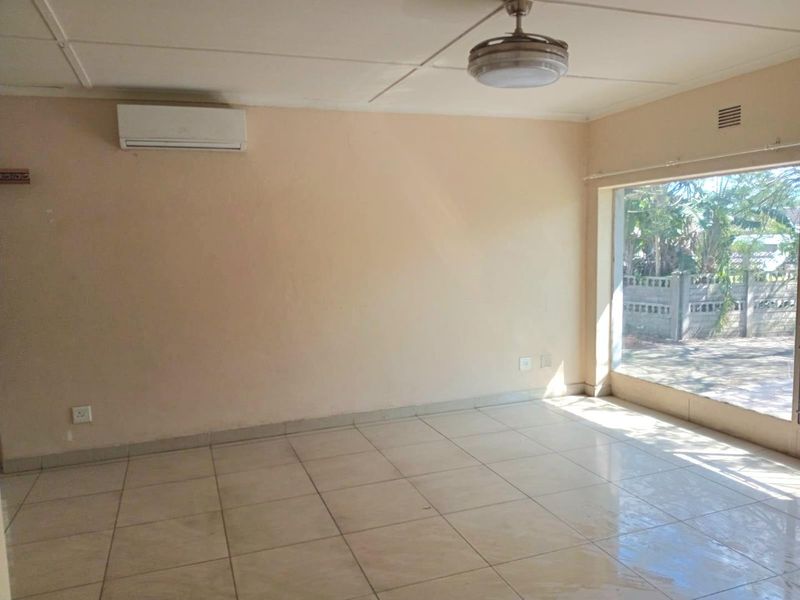 An empty living room featuring tiled flooring, a ceiling fan, and a wall-mounted air conditioning unit with large windows providing natural light.