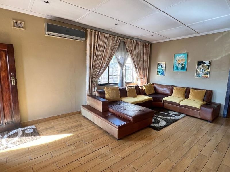 A spacious living room featuring tiled flooring, a wall-mounted air conditioning unit, and natural light through a window with security bars.