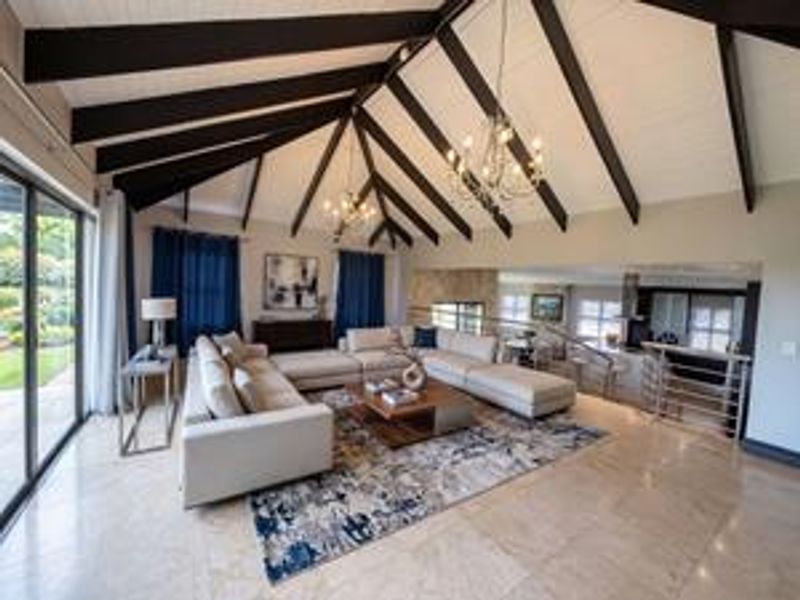 Spacious open-plan living area featuring vaulted ceilings with exposed timber beams, stone tile flooring, and large sliding glass doors leading to the outdoors.