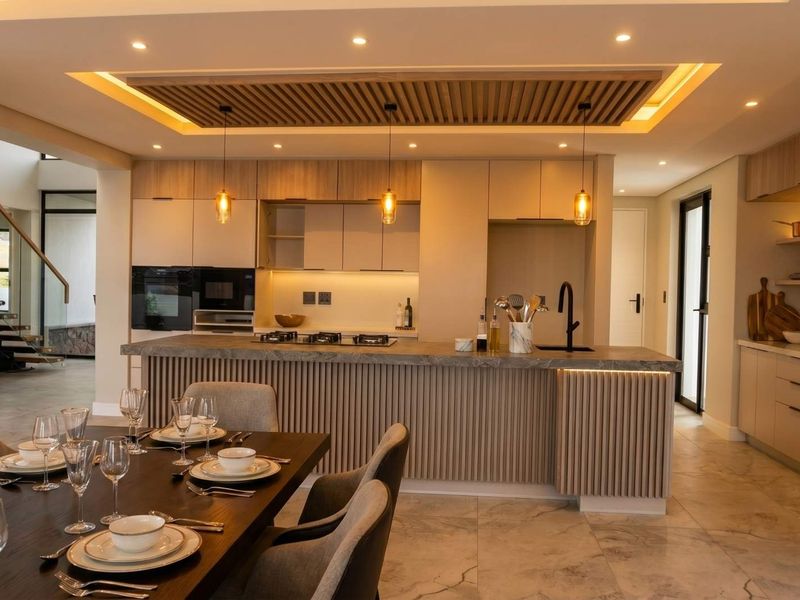 Modern open-plan kitchen and dining area featuring a timber-slat ceiling detail, stone-topped island, integrated appliances, and a sleek staircase with glass balustrade.